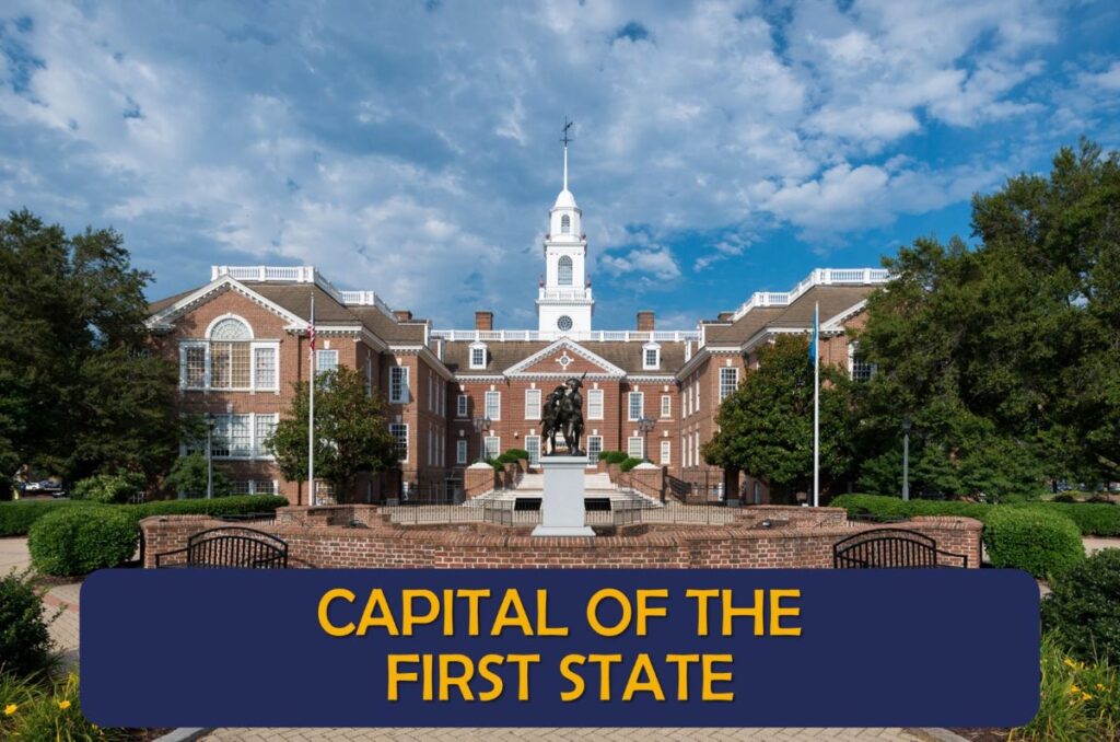 A historical brick building with a central clock tower and statue in front, framed by flags and greenery under a blue sky with scattered clouds. A banner at the bottom reads, "CAPITAL OF THE FIRST STATE.